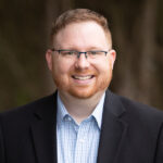 Smiling man with short red hair and beard wearing glasses, a light blue checkered shirt, and a black blazer outdoors. | Bryan Turner