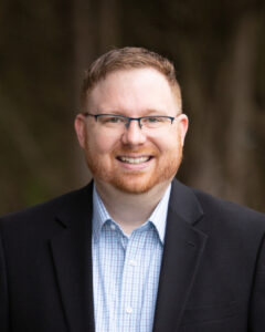 Smiling man with short red hair and beard wearing glasses, a light blue checkered shirt, and a black blazer outdoors. | Bryan Turner