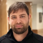 Portrait of a man with short dark hair and beard, wearing a black zip-up jacket, indoors with wooden columns in the background. | Bryce Woerner
