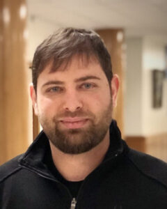 Portrait of a man with short dark hair and beard, wearing a black zip-up jacket, indoors with wooden columns in the background. | Bryce Woerner