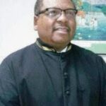 Smiling man in a black clerical cassock wearing glasses, indoors with a colorful wall art in the background. | Donald King