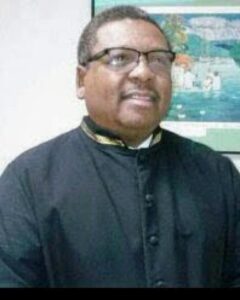 Smiling man in a black clerical cassock wearing glasses, indoors with a colorful wall art in the background. | Donald King