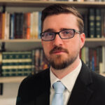 Head-and-shoulders portrait of a man with glasses, a beard, wearing a black suit and light blue tie, standing in front of a bookshelf. | Jason Quintern