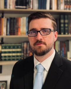 Jason Quintern | Jason Quintern | Cornerstone College & Seminary | Catalog Head-and-shoulders portrait of a man with glasses, a beard, wearing a black suit and light blue tie, standing in front of a bookshelf. | Jason Quintern