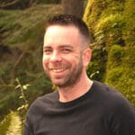 Man with short dark hair and trimmed beard smiling at camera outdoors with greenery behind him.