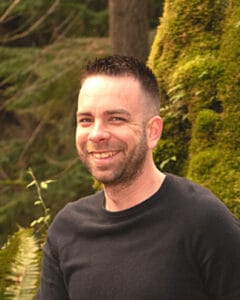Man with short dark hair and trimmed beard smiling at camera outdoors with greenery behind him. | Logan Castle