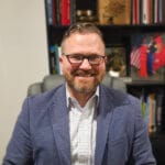 Smiling man with glasses and a beard wearing a blue blazer, seated in an office with bookshelves behind him.