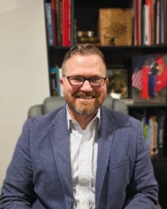 Smiling man with glasses and a beard wearing a blue blazer, seated in an office with bookshelves behind him. | Michael Hensley