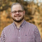 Smiling man with glasses and a beard in a red-and-white plaid shirt, outdoors with fall foliage.