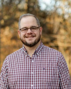 Smiling man with glasses and a beard in a red-and-white plaid shirt, outdoors with fall foliage. | Russell Threet
