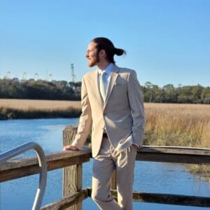 Man in a light tan suit leaning on a wooden railing beside a blue lake on a sunny day, looking to the left in profile. | Samuel Rhoades
