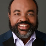 Smiling man with a dark beard and suit jacket looking at the camera. | Shawn Dandridge