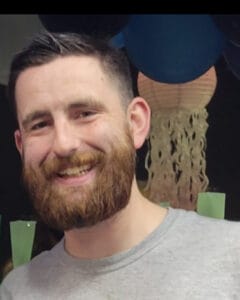 Smiling man with a brown beard and short dark hair, wearing a gray T-shirt. | Dillon Pegram
