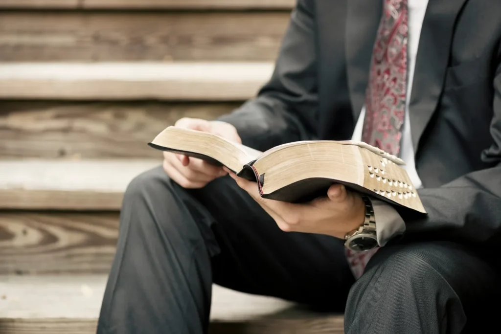Person in a suit reading a large, studded book while sitting on wooden steps outside desaturated tone.