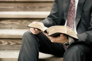 Undergraduate Certificate of Biblical Studies | Certificate of Biblical Studies – 2026-2027 | Cornerstone College & Seminary | Catalog Person in a suit reading a large, studded book while sitting on wooden steps outside desaturated tone. | Certificate of Biblical Studies – 2026-2027