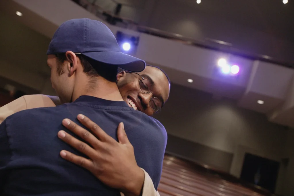 Two people hug warmly in a large indoor venue, one wearing a blue cap, stage lights in the background
