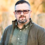 Portrait of a middle-aged man with short dark hair, beard, and glasses wearing a green button-up shirt and olive jacket outdoors.