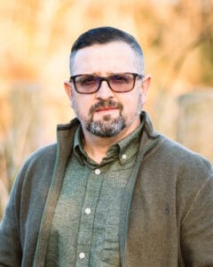 Portrait of a middle-aged man with short dark hair, beard, and glasses wearing a green button-up shirt and olive jacket outdoors. | Patrick Allen
