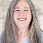 Close-up of a smiling woman with long gray hair, wearing a pale blue shirt, against a blurred brick wall background.
