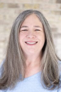 Close-up of a smiling woman with long gray hair, wearing a pale blue shirt, against a blurred brick wall background. | Jennifer Hanes